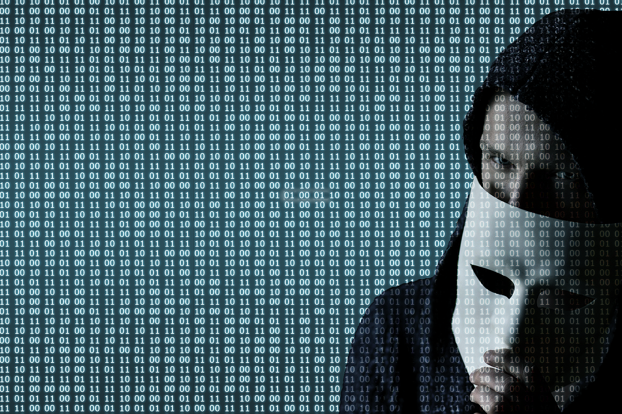 Picture of a hacker wearing a hoodie and holding a mask and the screen shows binary code all over it.